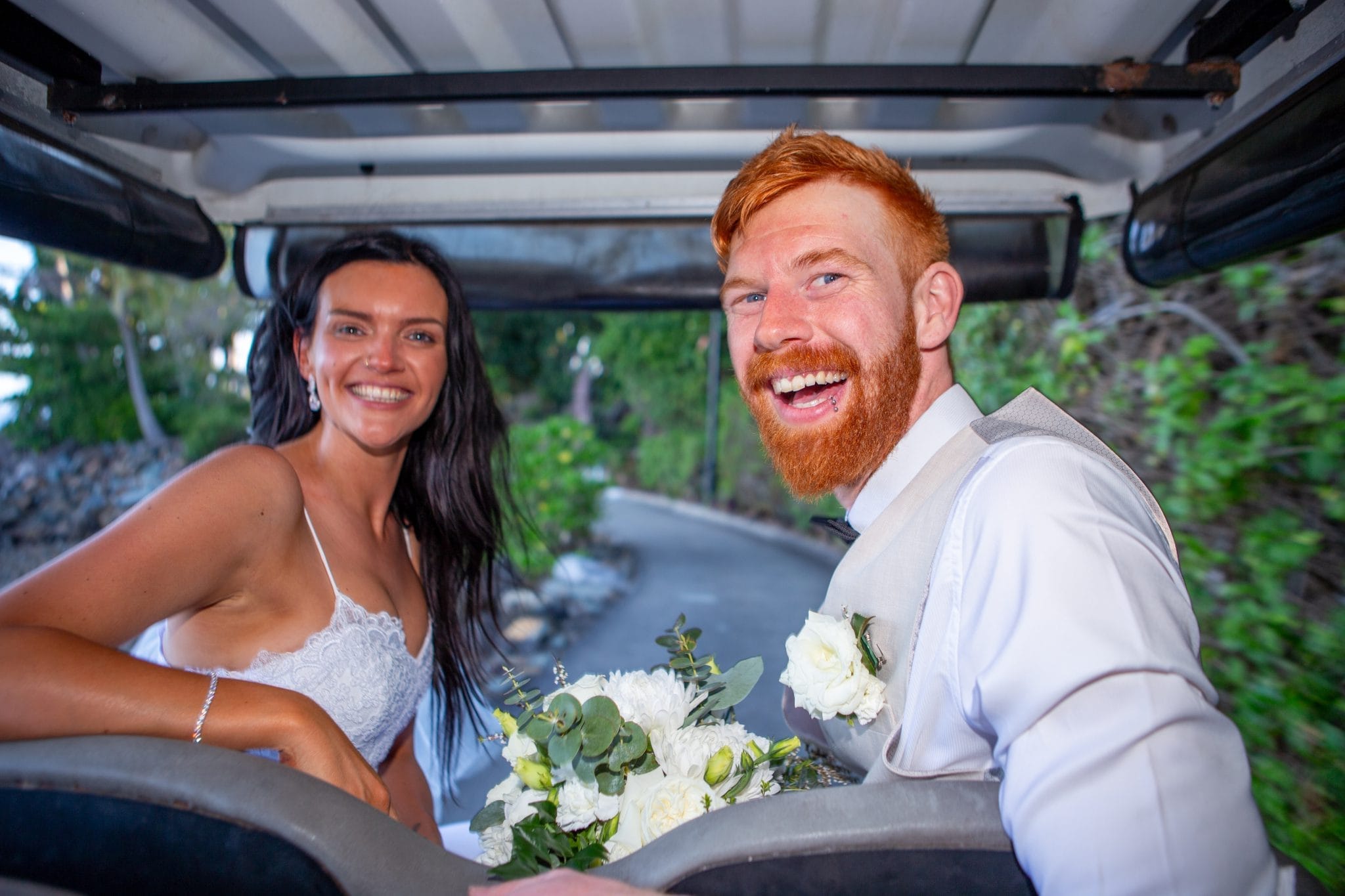 Bride and groom sitting on the back of a golf cart smiling together.