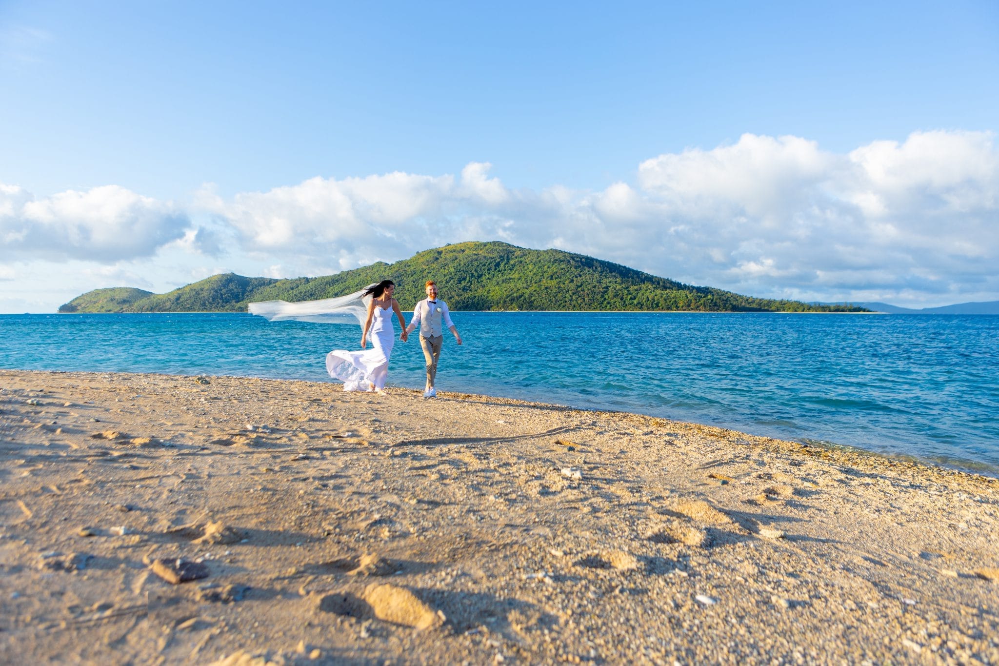 Bride and groom walking along a sandy beach in front of blue ocean